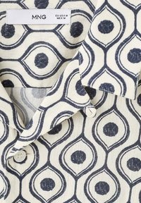 Camisa de manga corta con un patrón geométrico en azul marino y crema. Presenta un collar puntiagudo, botones y una textura de tela suave.