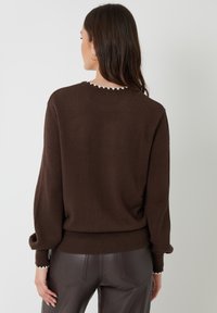 Brauner Strickpullover mit einem gewelltem weißen Ausschnitt, lockerer Passform und geripptem Saum. Kombiniert mit dunkelbraunen, eng anliegenden Hosen.