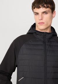 Schwarze Jacke mit einem Reißverschluss an der Vorderseite, Kapuze und geripptem Textil am oberen Körper. Verfügt über kontrastierendes Material an den Schultern und schlanke Taschen-details.
