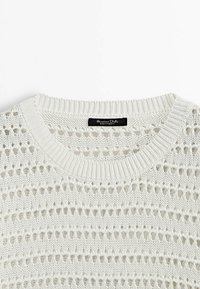 Pull blanc en tricot avec des rayures horizontales en ajouré et un col rond côtelé, avec une étiquette noire cousue à l'intérieur du col.