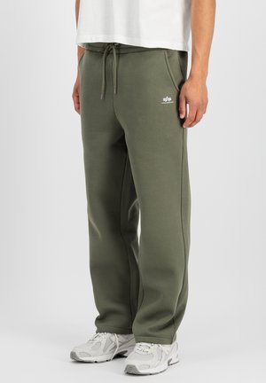 Man wearing olive green sweatpants with drawstring and white logo on thigh, white sneakers, and white shirt standing against plain background.