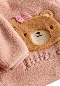 Fleece rozā apģērbs ar brūnu lāča seju un rozā loku, un uz apakšējās daļas izbrodēts vārds "mazais" violetā krāsā.