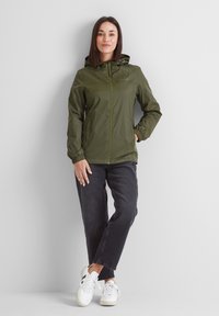 Green waterproof jacket features a front zipper, adjustable hood, and elastic cuffs, paired with black jeans and white sneakers.