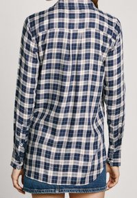 Person klädd i en marinblå och vit rutig långärmad skjorta med knäppta manschetter, kombinerad med en blå denimkjol, sedd bakifrån.