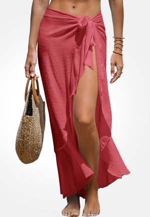 Langer, pinkfarbener Wickelrock mit Bindeschluss, der einen hohen Schlitz und einen gerafften Saum aufweist. Model hält eine geflochtene Tragetasche, barfuß.