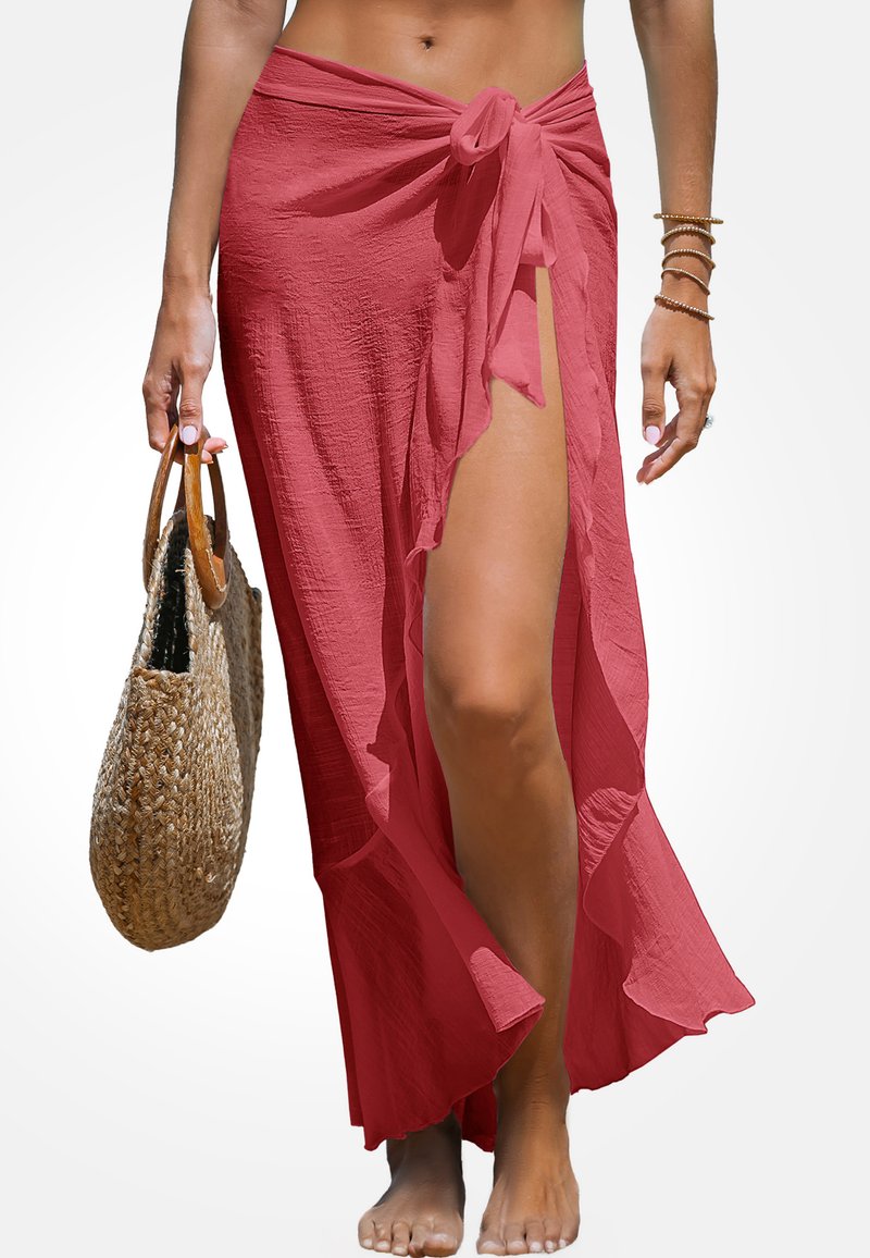Długa, różowa spódnica owijana z zawiązywaniem, z wysokim rozcięciem i falbaną na brzegu. Modelka trzyma plecioną torbę, bosa.