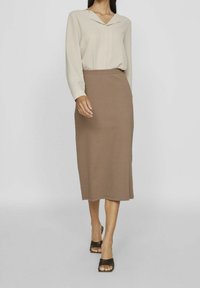 Donna che indossa un top beige a maniche lunghe, una gonna midi taupe e sandali con tacco aperti neri, mentre cammina su uno sfondo neutro.