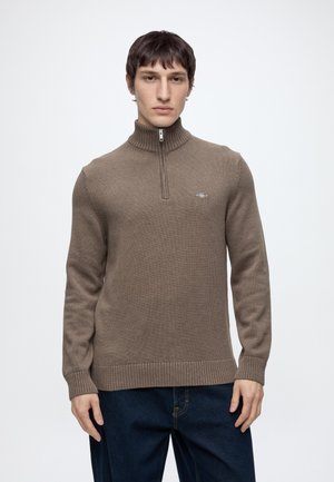 Junger Mann trägt einen braunen Halb-Reißverschluss-Pullover und dunkelblaue Jeans, steht vor einem einfarbigen hellen Hintergrund.