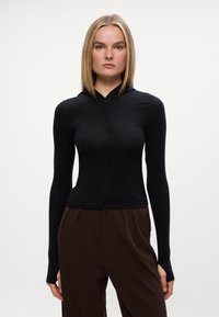 Sort langærmet top med lynlås i halsen, tætsiddende design og blød tekstur, kombineret med løse brune bukser. Minimalistisk stil.