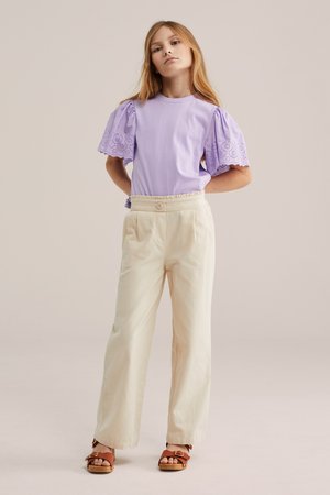 Młoda dziewczyna stojąca z rękami za plecami, ubrana w lawendowy top z bufkami, beżowe spodnie o szerokich nogawkach i brązowe sandały.