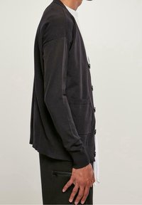 Svart jersey cardigan med knappar, med en avslappnad passform, ribbade ärmslut och sidofickor. Bärs över en vit t-shirt och svarta byxor.