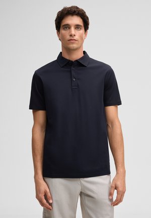 Man die recht vooruit kijkt en een zwart poloshirt met korte mouwen en lichtgekleurde broek draagt tegen een effen achtergrond.