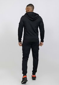 Survêtement de sport noir comprenant un sweat à capuche et un pantalon cargo. Tissu doux, design ajusté, avec des poches sur le pantalon et des accents de couleur sur les chaussures.