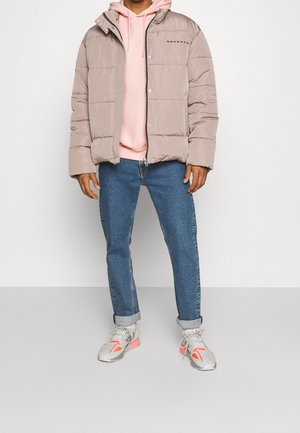 Hombre con chaqueta acolchada beige sobre sudadera con capucha rosa, vaqueros azules remangados y zapatillas blancas con detalles naranjas, de pie sobre un fondo liso.