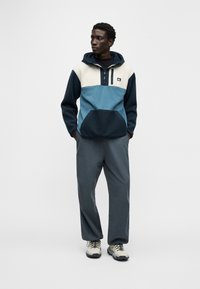 Jauns vīrietis stāv ar rokām kabatās, valkājot zilas, krēmkrāsas un tumši zila krāsas kapucei piederošu flīsa jaku, vaļīgas pelēkas bikses un bēšas pārgājienu kurpes.