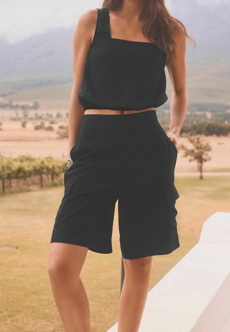 Femme portant un haut noir sans manches et un short noir jusqu'aux genoux, debout en plein air les mains dans les poches, paysage naturel flou en arrière-plan.