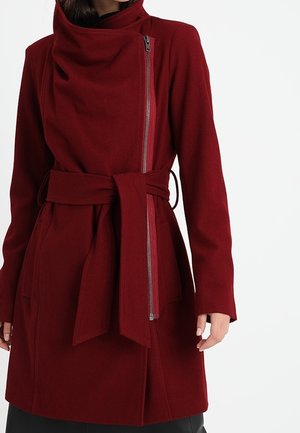 Kobieta ubrana w ciemnoczerwony płaszcz z paskiem, z asymetrycznym srebrnym zamkiem błyskawicznym i wysokim kołnierzem, stojąca na jednolitym białym tle.