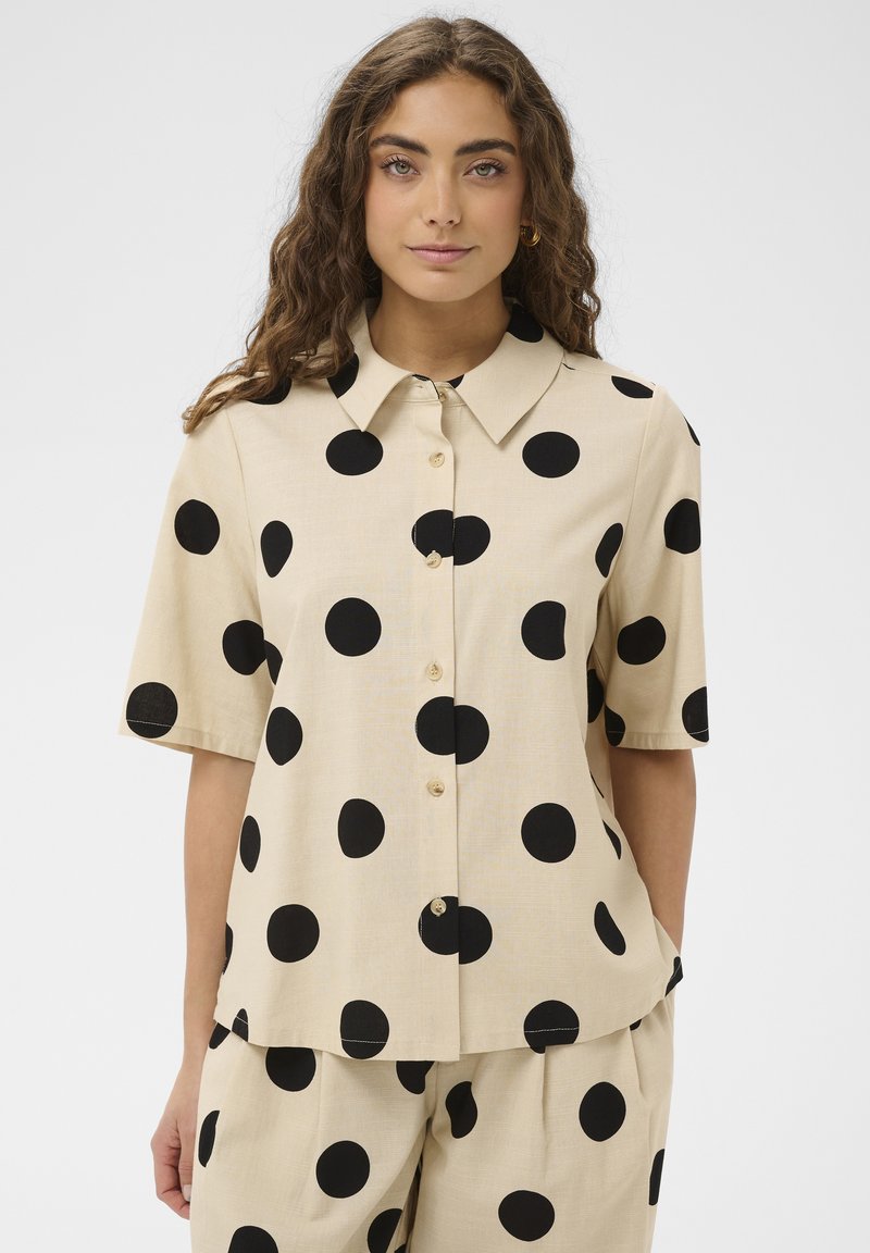 Femme aux longs cheveux bouclés portant une chemise beige à manches courtes avec boutons et un pantalon assorti à grands pois noirs sur un fond uni.