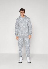 Pelēks kapučjakas un sporta bikses komplekts ar abstraktām baltām rakstu zīmēm, izgatavots no mīksta auduma, ar kenguru kabatu un ribotām aprocēm.