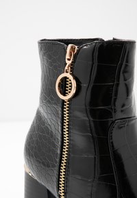 Svart patenterad ankelsko med krokodilmönster, med en gyllene dragkedja och rund dragflik. Glansig yta med klack i blockmodell.