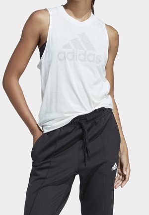 Primo piano di una persona che indossa un top bianco senza maniche Adidas e pantaloni neri Adidas con coulisse, con le mani sui fianchi.