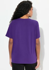 Mujer con cabello largo y rizado negro, vestida con una camisa corta de manga púrpura sencilla y pantalones negros, de espaldas contra un fondo blanco.