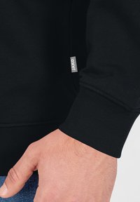 Sweatshirt noir avec des poignets et un ourlet côtelés, présente une petite étiquette grise "JAKO" sur le côté. Tissu texturé, design ajusté.
