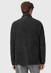 Homme de dos portant une veste en daim noir avec des poignets boutonnés et un jean en denim gris clair, le tout sur un fond clair et uni.