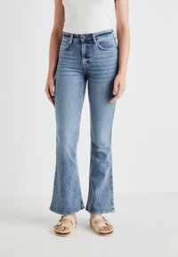 Γυναίκα που φοράει ανοιχτό μπλε ψηλόμεσα φαρδιά τζιν και μπεζ σανδάλια με δύο λουριά, στέκεται μπροστά σε λευκό φόντο.