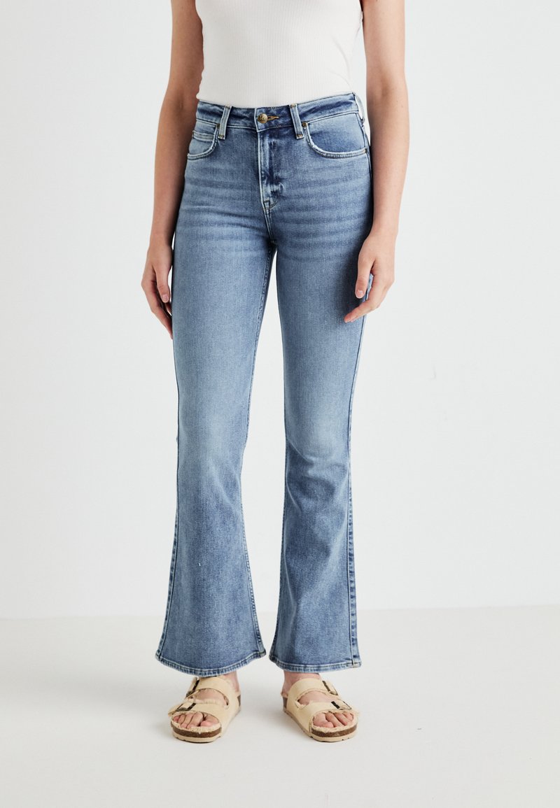 Γυναίκα που φοράει ανοιχτό μπλε ψηλόμεσα φαρδιά τζιν και μπεζ σανδάλια με δύο λουριά, στέκεται μπροστά σε λευκό φόντο.