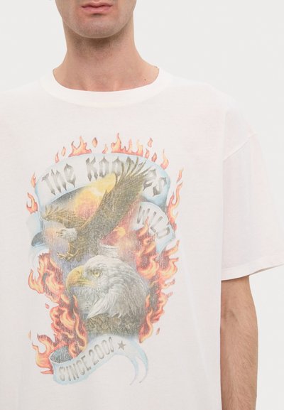 Camiseta blanca de algodón con un gráfico multicolor de águilas, llamas y una cinta con "Desde 2008" en letras en negrita. Corte relajado.