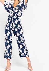 Žena v námořnickém modrém květinovém overalu s širokými nohavicemi a volnými rukávy, v béžových sandálech na blokovém podpatku, stojí s rukama vztyčenýma.