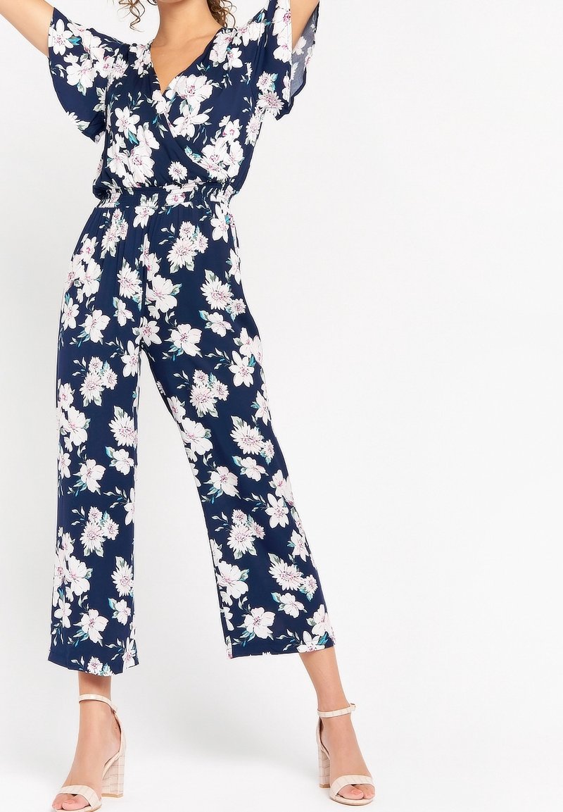 Žena v námořnickém modrém květinovém overalu s širokými nohavicemi a volnými rukávy, v béžových sandálech na blokovém podpatku, stojí s rukama vztyčenýma.