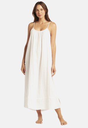 Una mujer luce un vestido blanco, suelto, sin mangas, hasta el tobillo con tirantes finos sobre un fondo claro y liso.
