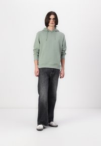Felpa di cotone verde menta con tasca frontale e cappuccio regolabile, abbinata a jeans larghi grigio scuro e sneakers grigie.