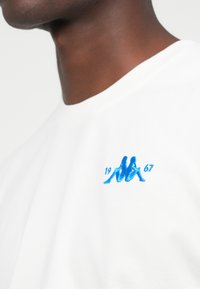 Nahaufnahme einer Person, die einen weißen Sweatshirt mit einem blauen gestickten Logo trägt, das zwei Rücken an Rücken sitzende Figuren und den Text "1967" zeigt.