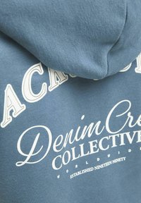 Felpa blu con branding in bianco. Il materiale sembra morbido e con una texture liscia. Il design include un cappuccio e lettering prominente.