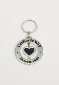 Srebrni okrugli privjesak za ključeve s crnim srcem u središtu i ugraviranim tekstom "Carhartt WIP Detroit 1989" oko rubova.