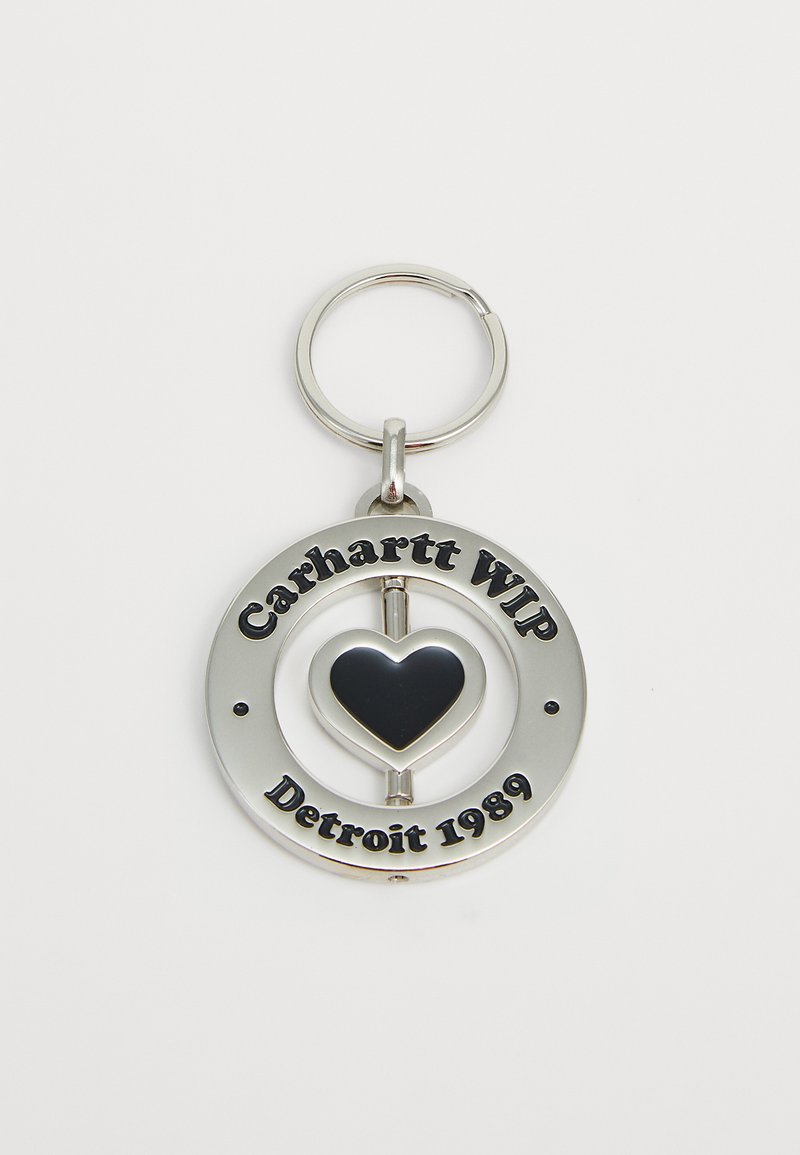 Srebrni okrugli privjesak za ključeve s crnim srcem u središtu i ugraviranim tekstom "Carhartt WIP Detroit 1989" oko rubova.