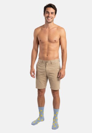 Kaki korte broek met voorzakken, blauwe sokken met gele eendjespatronen, gedragen door een man met kort haar, staand tegen een effen achtergrond.