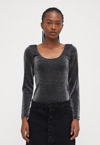 Svart langermet bodysuit i et skinnende, teksturert stoff med dyp U-hals, iført en svart knappet skjørt som har en tettsittende design.