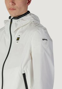 Giacca bianca con cappuccio realizzata in materiale leggero, dotata di una cerniera nera, colletto con strisce nere e bianche e un patch con logo sul braccio.