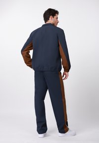 Μπλε ναυτικό και καφέ φόρμα με μπουφάν με φερμουάρ, ελαστικές μανσέτες και παντελόνι με ρίγες στο πλάι και άνετη εφαρμογή. Άσπρα αθλητικά παπούτσια.