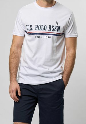 Mężczyzna w białej koszulce U.S. Polo Assn. z logo i napisem, w połączeniu z granatowymi szortami, stojący na jednolitym tle.