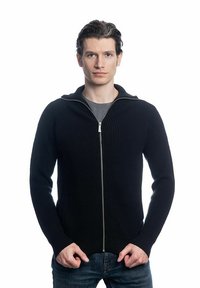 Pull en maille côtelée noir à fermeture éclair avec un col montant, doté d'une fermeture éclair en argent et de poches latérales. Porté par-dessus un T-shirt gris et un jean bleu.