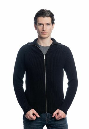 Pull en maille côtelée noir à fermeture éclair avec un col montant, doté d'une fermeture éclair en argent et de poches latérales. Porté par-dessus un T-shirt gris et un jean bleu.