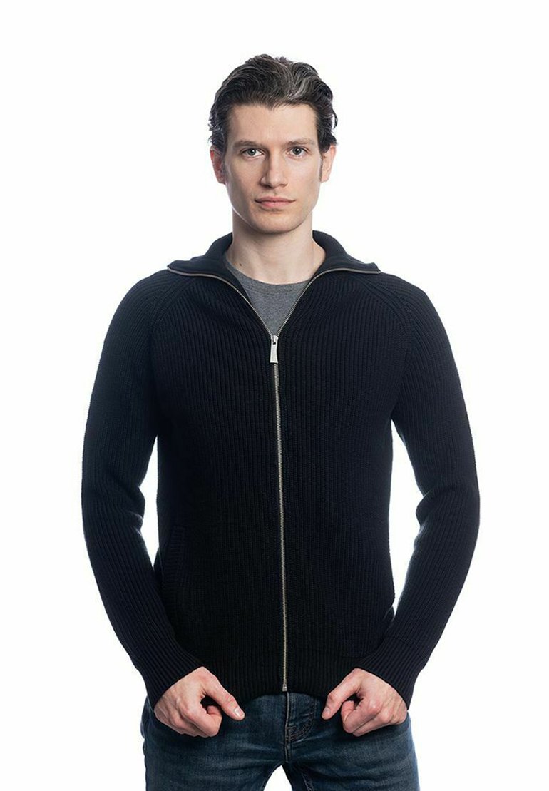 Pull en maille côtelée noir à fermeture éclair avec un col montant, doté d'une fermeture éclair en argent et de poches latérales. Porté par-dessus un T-shirt gris et un jean bleu.