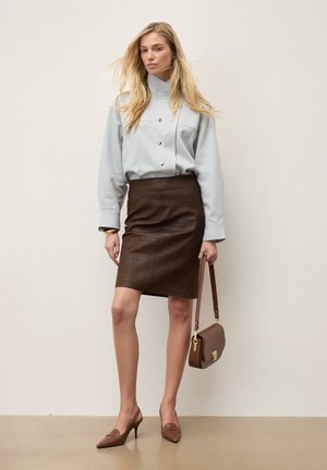 Frau trägt eine hellgraue, geknöpfte Bluse und einen dunkelbraunen ledernen kniehohen Rock, hält eine braune Handtasche und trägt passende braune Absätze.