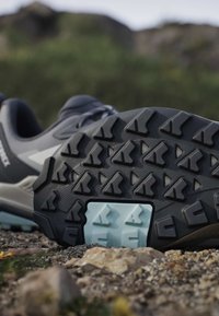 Gros plan de la semelle noire d'une chaussure de randonnée avec un détail de traction bleu reposant sur un sol rocheux en extérieur.