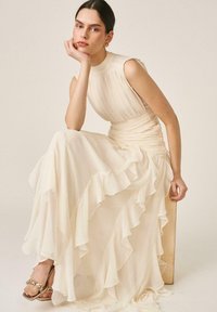 Mujer con cabello oscuro recogido, que lleva un vestido crema plisado sin mangas con falda con volantes, sentada apoyando la barbilla en la mano contra un fondo liso.